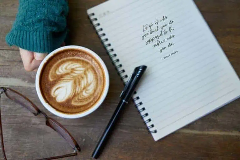 a cozy flatlay featuring a hand holding a cup of coffee with latte art, a spiral notebook with a brené brown quote, a black pen, and a pair of glasses placed on a rustic wooden table