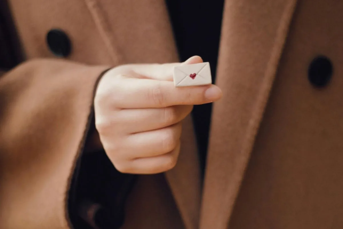 a hand holding a tiny envelope with a red heart symbol, wearing a brown coat