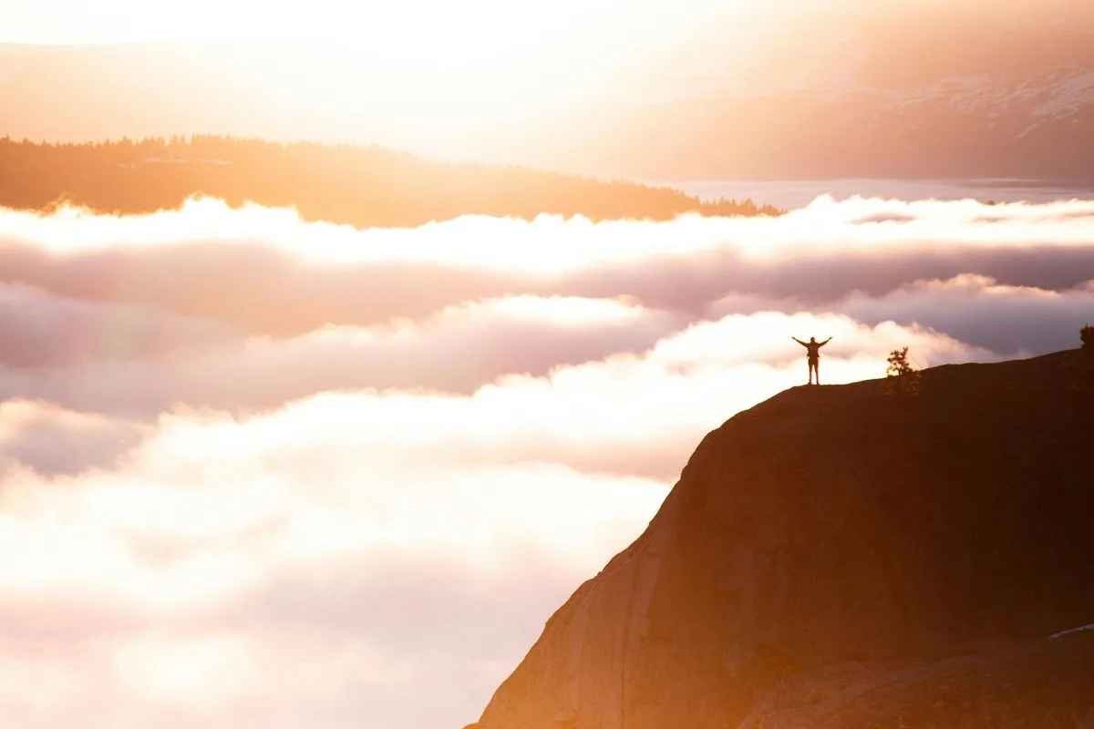 person standing triumphantly on a mountain peak surrounded by glowing clouds at sunrise