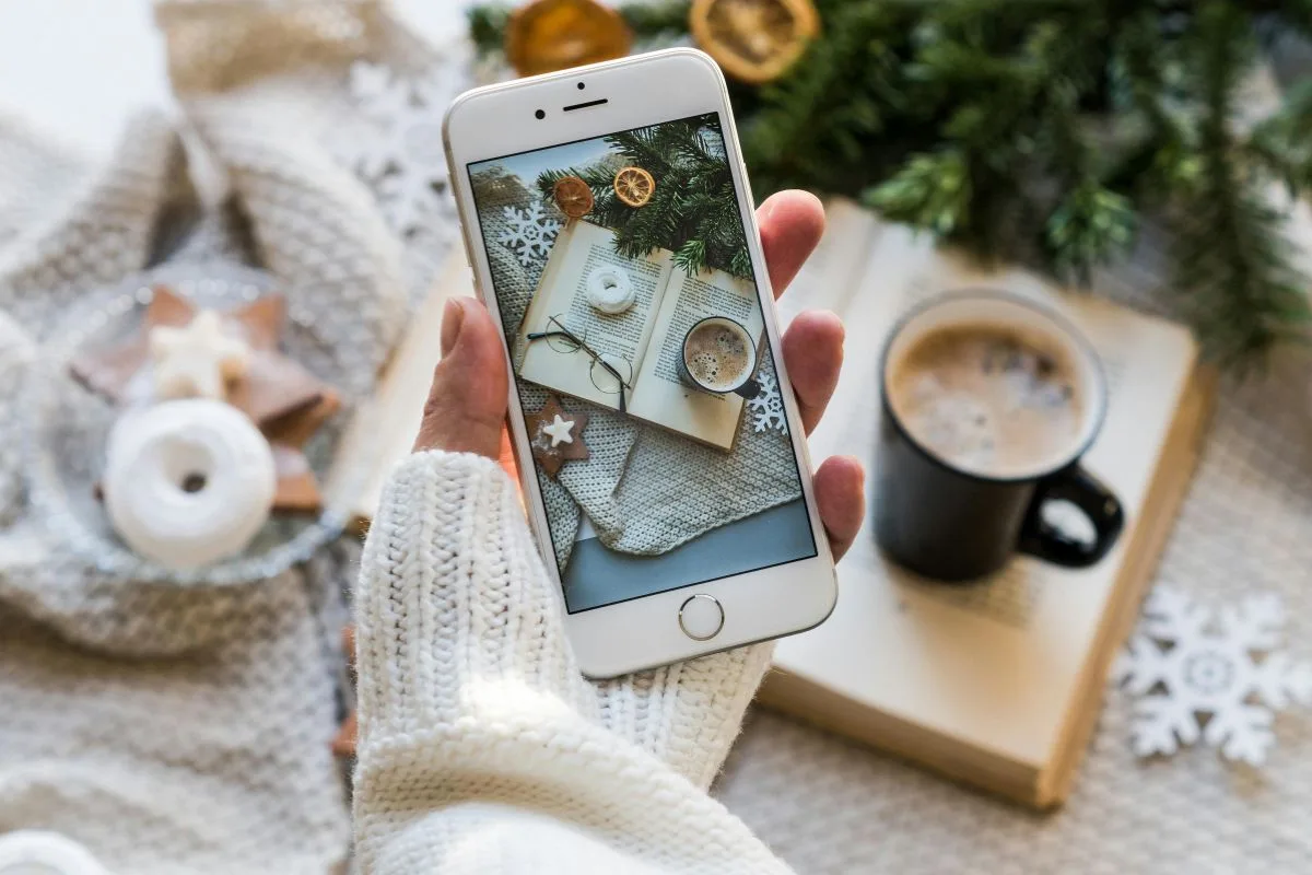 cozy Christmas flatlay with a hand holding a phone capturing a festive scene of coffee, cookies, a book, and winter decorations
