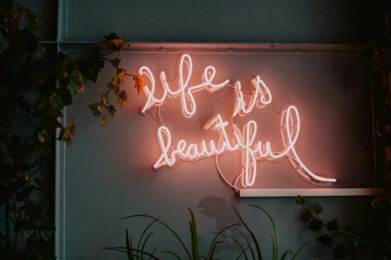 neon sign glowing with the words life is beautiful on a wall surrounded by green vines