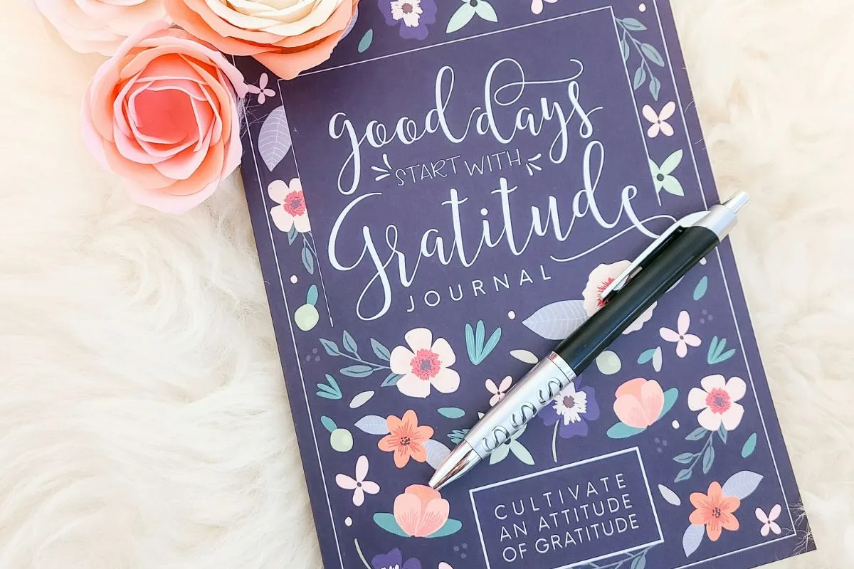 an open gratitude journal with floral illustrations, a pen, and pastel paper flowers placed on a soft white fur surface