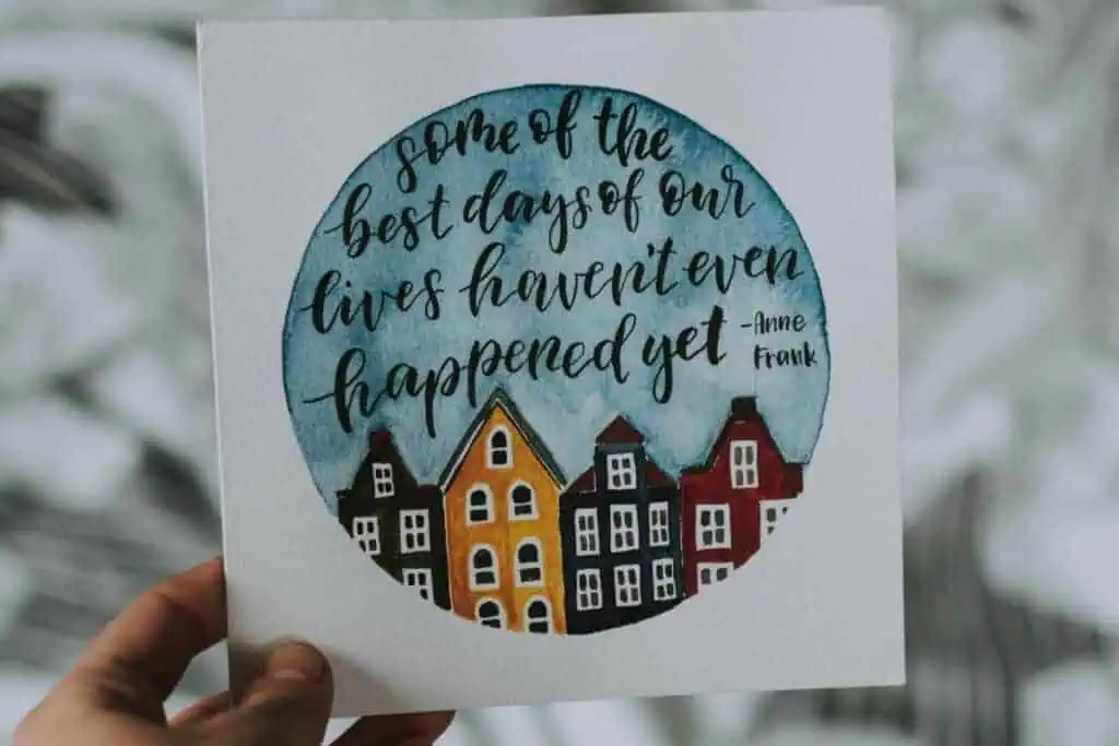 a hand holding a card with an inspirational quote by anne frank and watercolor houses in the background