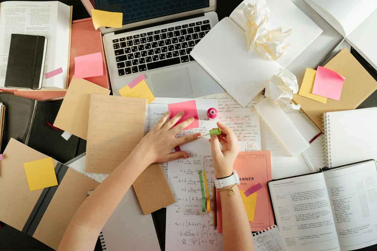 hands working amidst scattered books, notes, and a laptop — a chaotic yet focused hustle moment