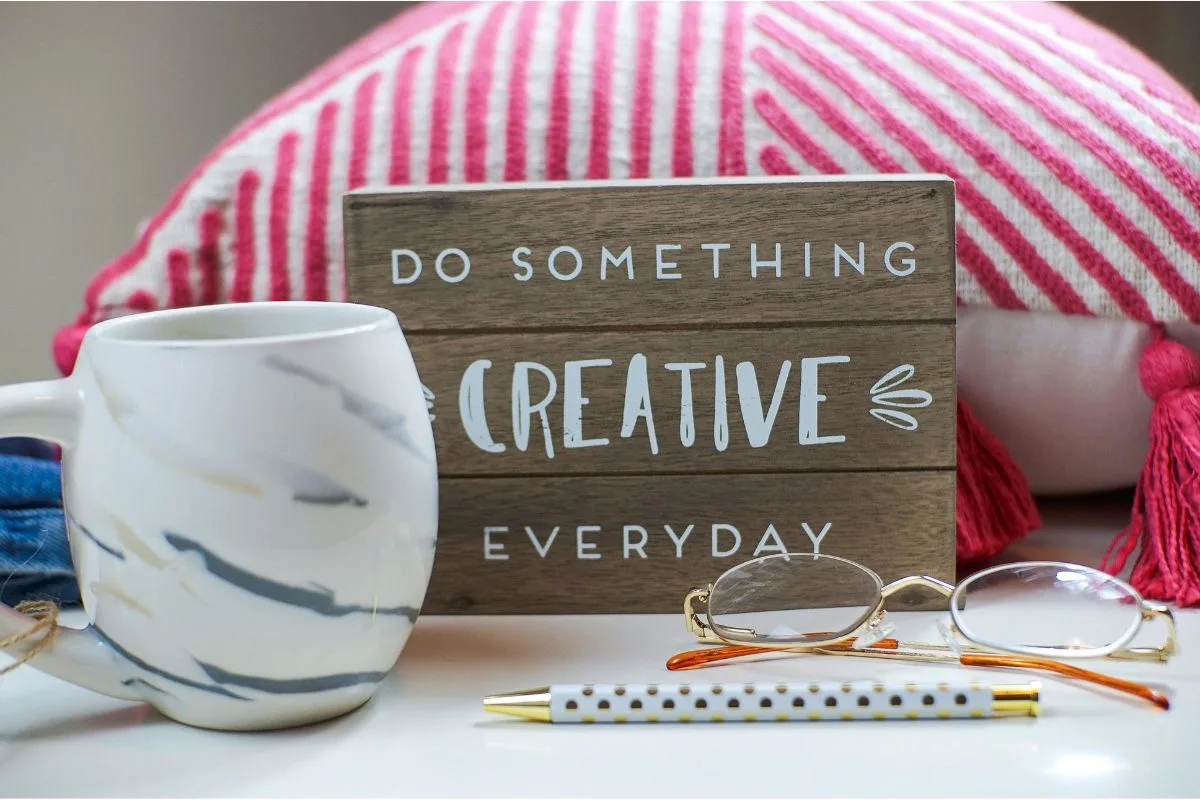 wooden sign with quote "do something creative everyday" placed on a desk with a coffee mug, pen, and glasses in front of a striped pillow