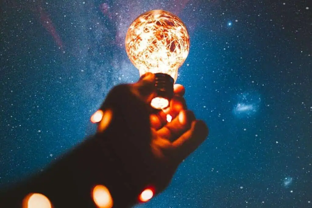 a hand holding a glowing light bulb wrapped in string lights against a star-filled night sky