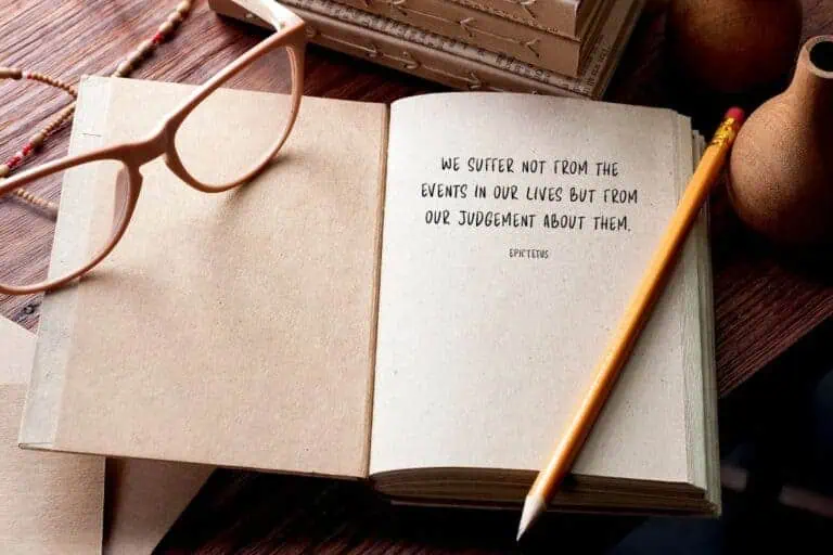open journal with an epictetus quote written on one page, placed beside beige glasses and a pencil on a wooden table