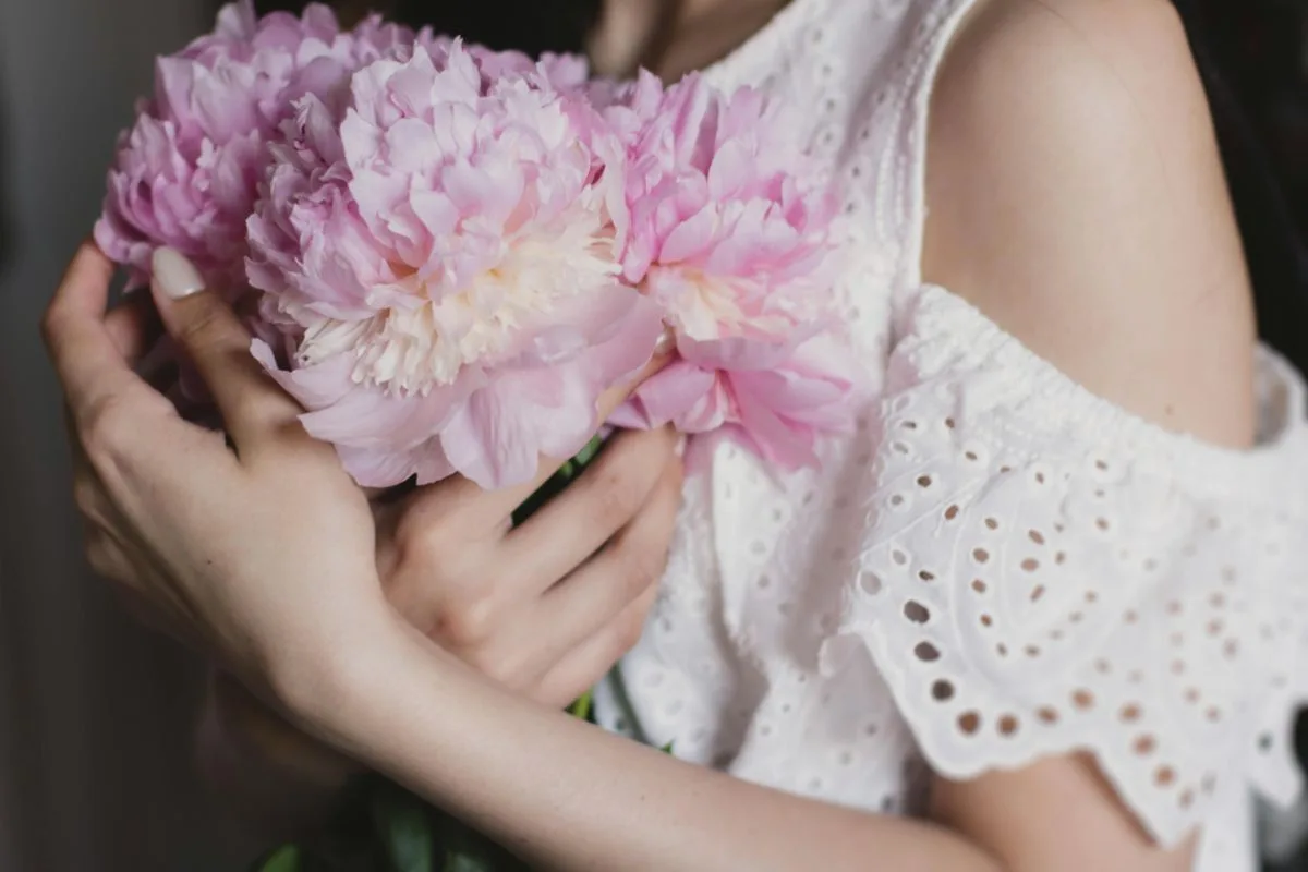 hands holding soft pink peonies in a white embroidered dress
