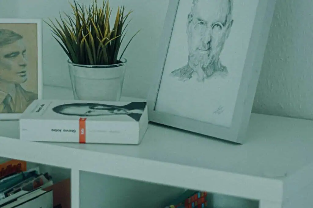 framed sketch of steve jobs placed on a white bookshelf beside books and a small potted plant