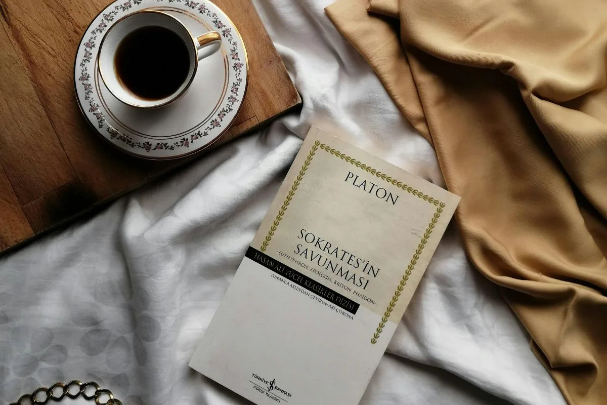 aesthetic flatlay featuring a turkish edition of plato’s apology of socrates with a cup of black coffee, gold silk fabric, and a delicate chain on a white bedsheet