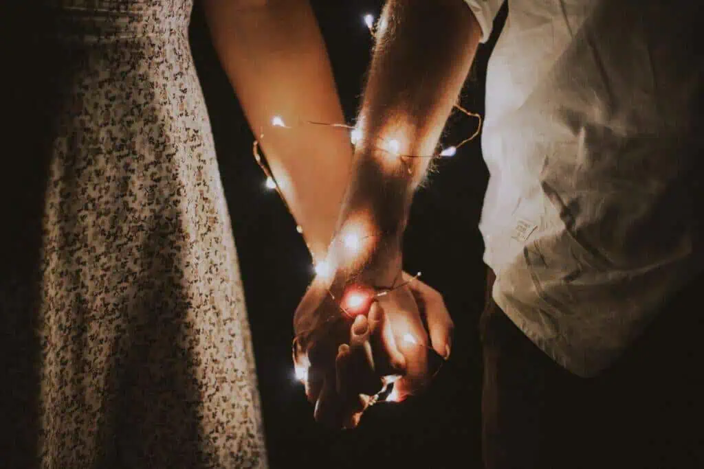 a close-up of a couple holding hands wrapped in delicate fairy lights against a dark background, capturing a quiet and intimate moment