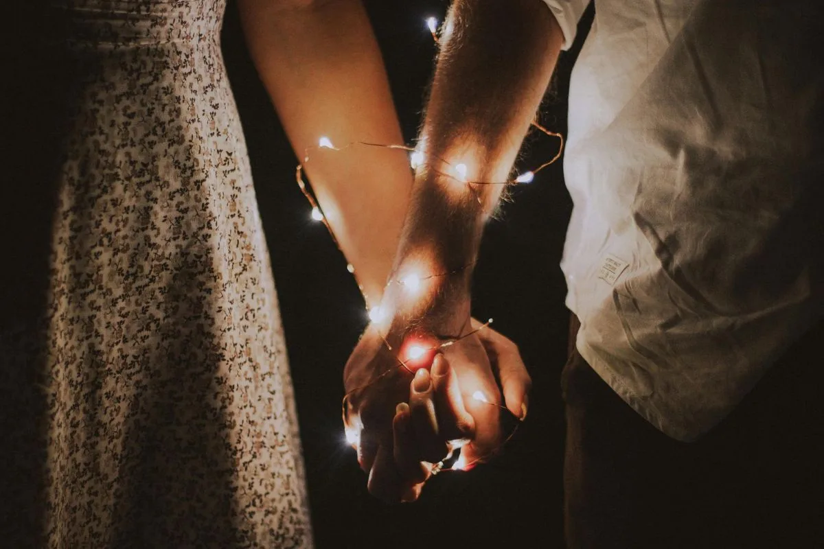 a close-up of a couple holding hands wrapped in delicate fairy lights against a dark background, capturing a quiet and intimate moment