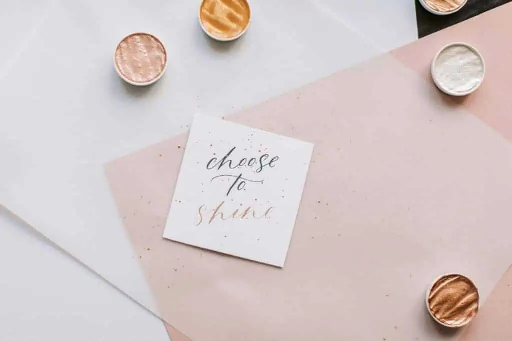 handwritten note with the words “choose to shine” placed on pastel paper with shimmer paint pots scattered around