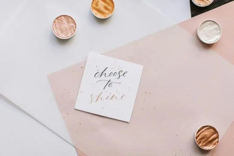 handwritten note with the words “choose to shine” placed on pastel paper with shimmer paint pots scattered around