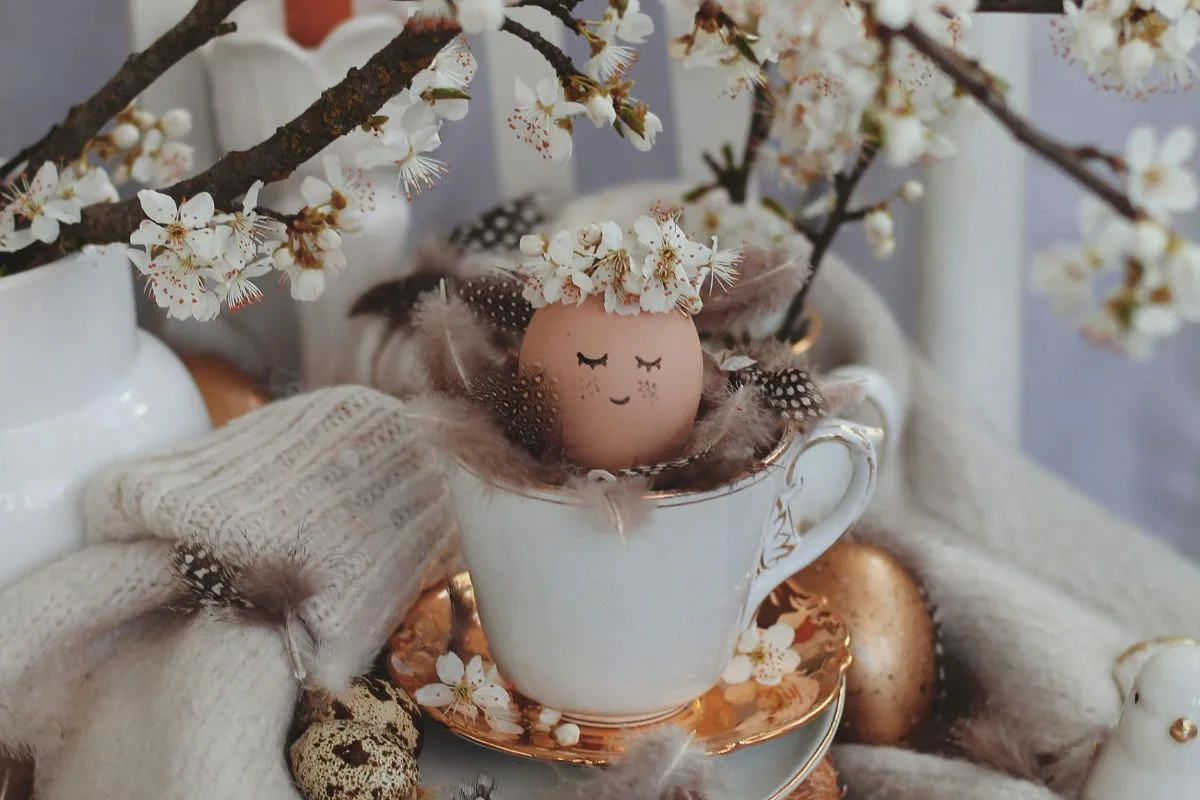 cozy spring scene with white cherry blossoms, a decorated egg with a flower crown resting in a teacup, surrounded by feathers, wool, and soft neutral tones