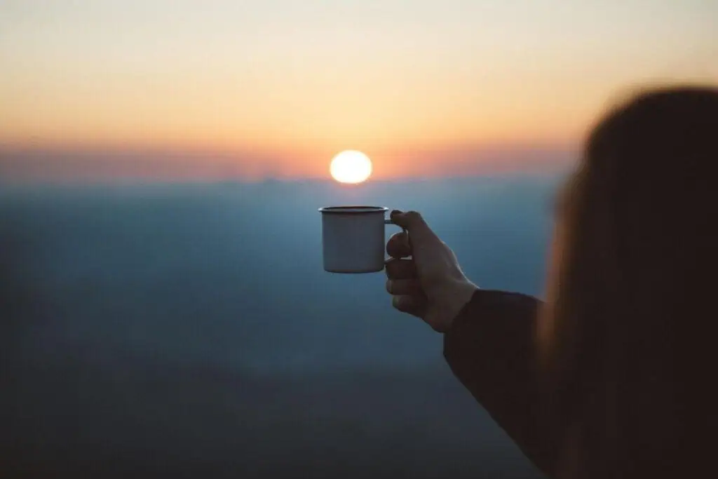hand holding a mug toward the rising sun over a soft, misty landscape