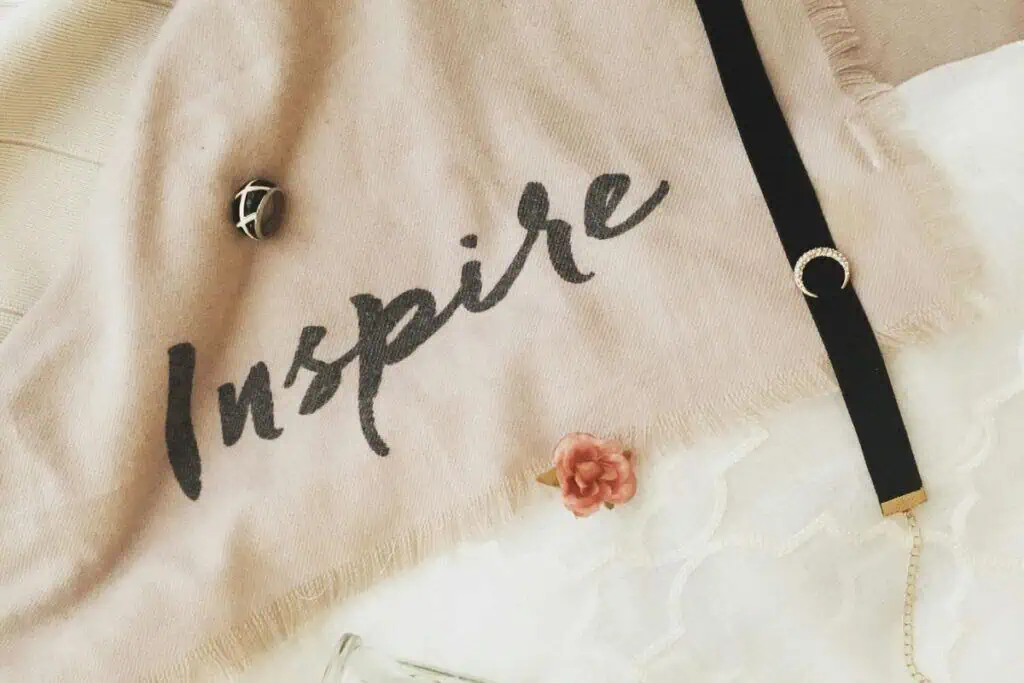 soft beige fabric with the word "inspire" written in black, styled with a ring, a bracelet, a tiny flower, and a glass vase on a white lace background