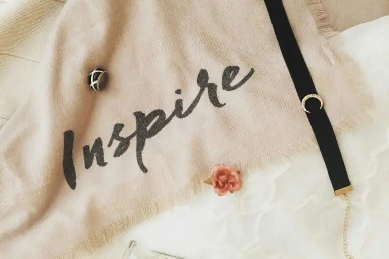 soft beige fabric with the word "inspire" written in black, styled with a ring, a bracelet, a tiny flower, and a glass vase on a white lace background