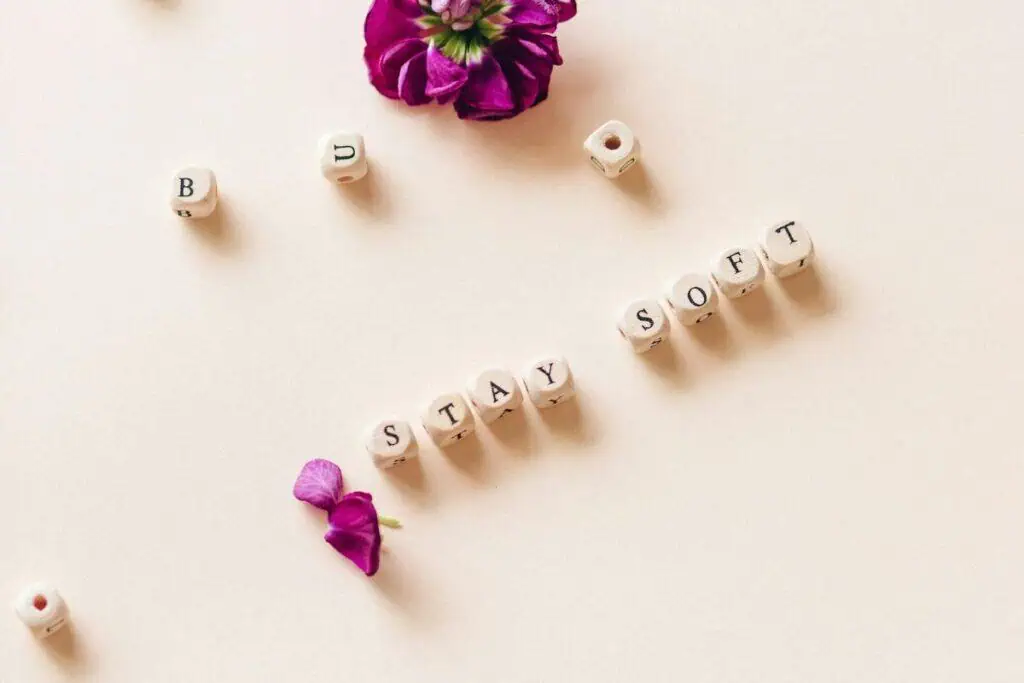 wooden letter beads spelling “stay soft” placed on a pastel surface with scattered purple flowers