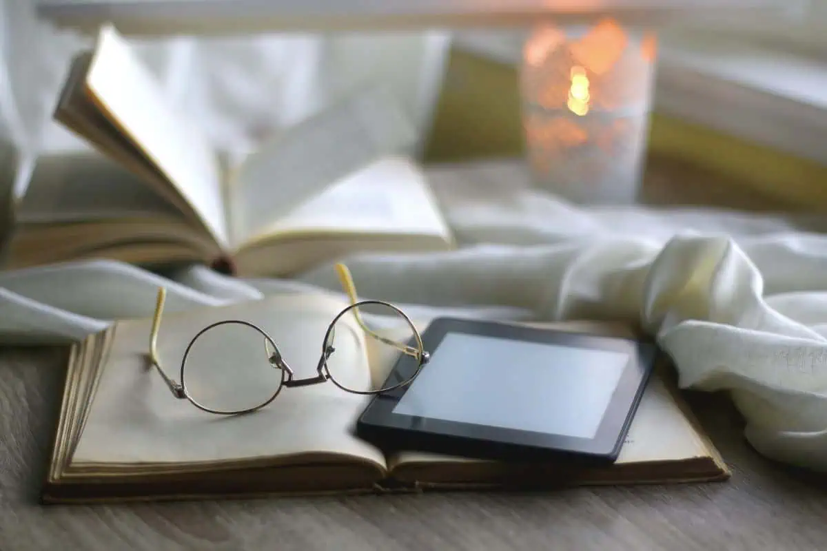 open book with reading glasses and e-reader on top, soft candlelight in background