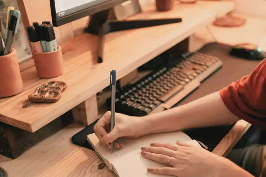 person writing in a notebook on a wooden desk with pens and computer keyboard nearby
