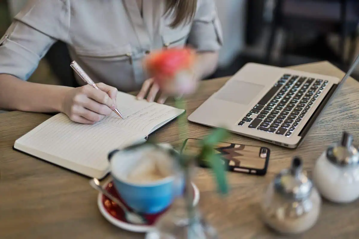woman writing in a notebook with a laptop, coffee cup, and phone on a wooden desk