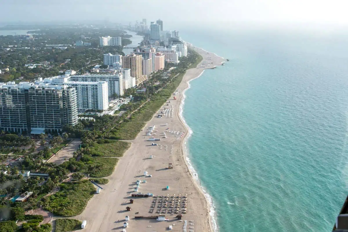 miami beach coastline with turquoise ocean, sandy shore, palm-lined buildings, and city skyline viewed from above