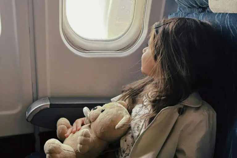 little girl sitting by airplane window holding a teddy bear and looking outside during flight, family travel with kids moment